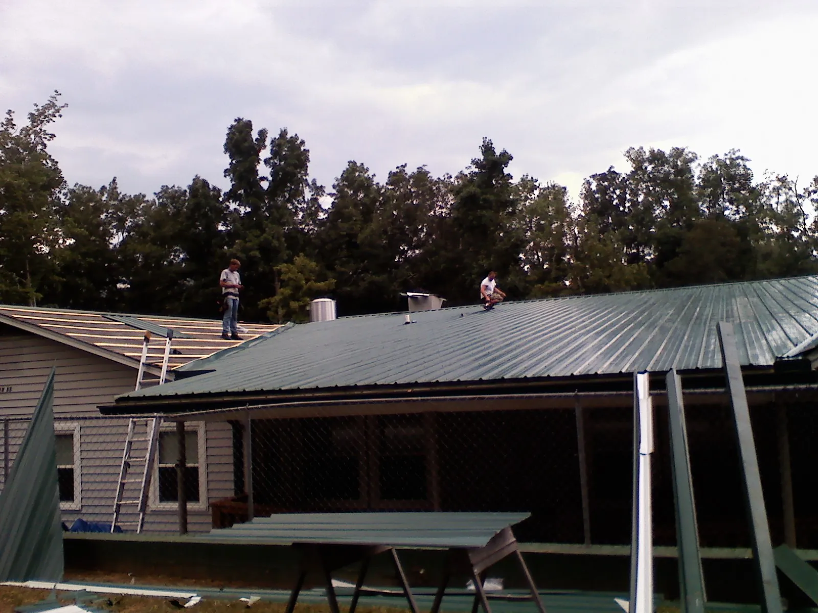 Two roofers installing green metal panels above a covered porch in Hancock